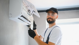 Entretien climatiseur : maintenance du filtre par un technicien. Technicien CVC souriant en casquette et lunettes inspecte un climatiseur mural. Il retire le filtre pour la maintenance et l'entretien.