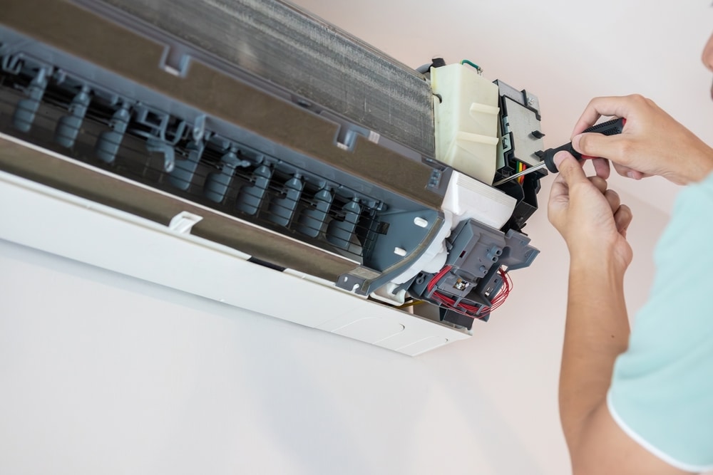 Réparation climatiseur : intervention technique et maintenance. Maintenance d'un climatiseur mural ouvert. Un technicien répare les circuits électriques internes avec un tournevis. Serpentins et ailettes visibles.